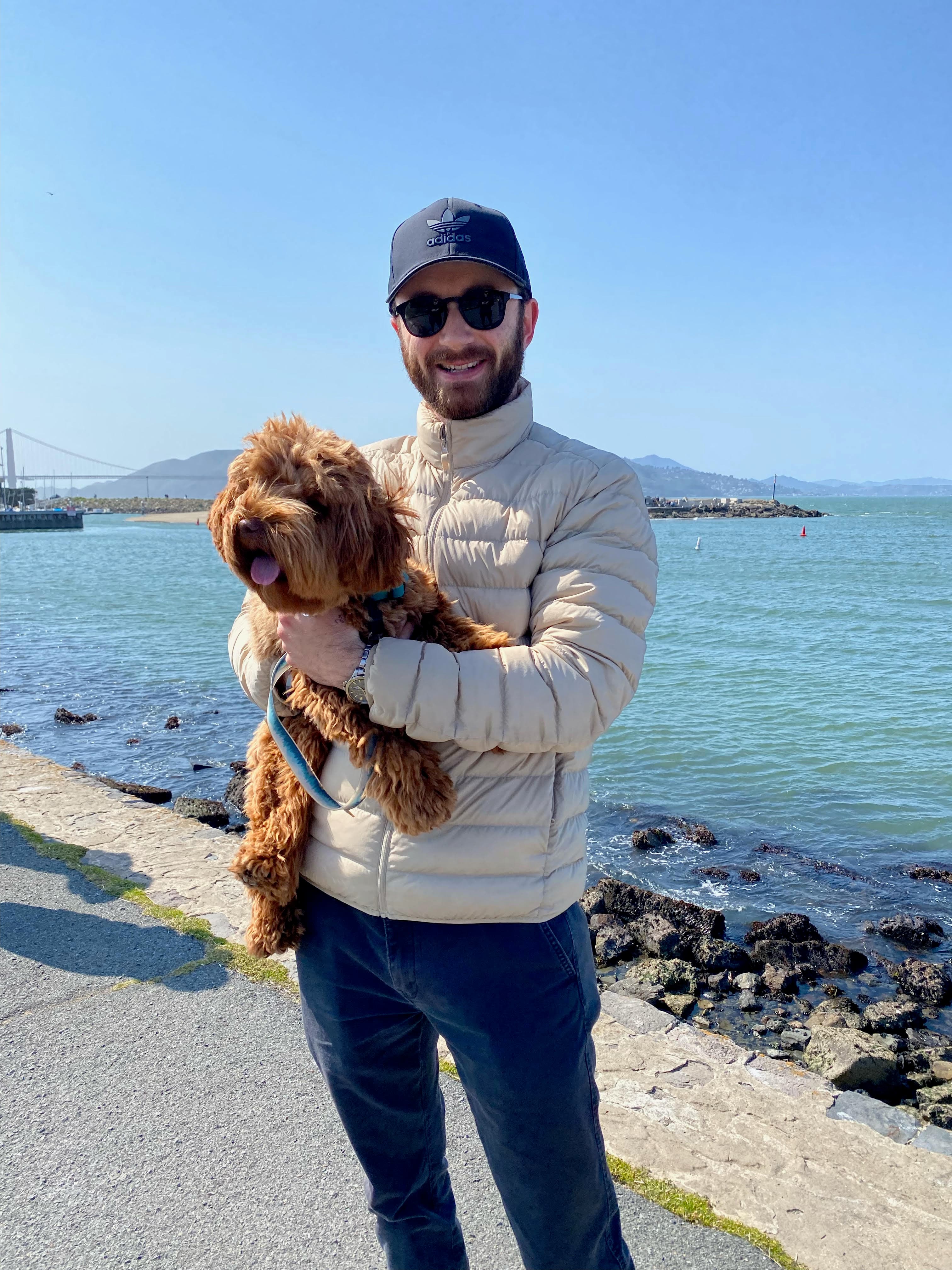 Hugo, co-founder of SF Pet Waste Removal, holding his labradoodle at the San Francisco waterfront with the Golden Gate Bridge in the background